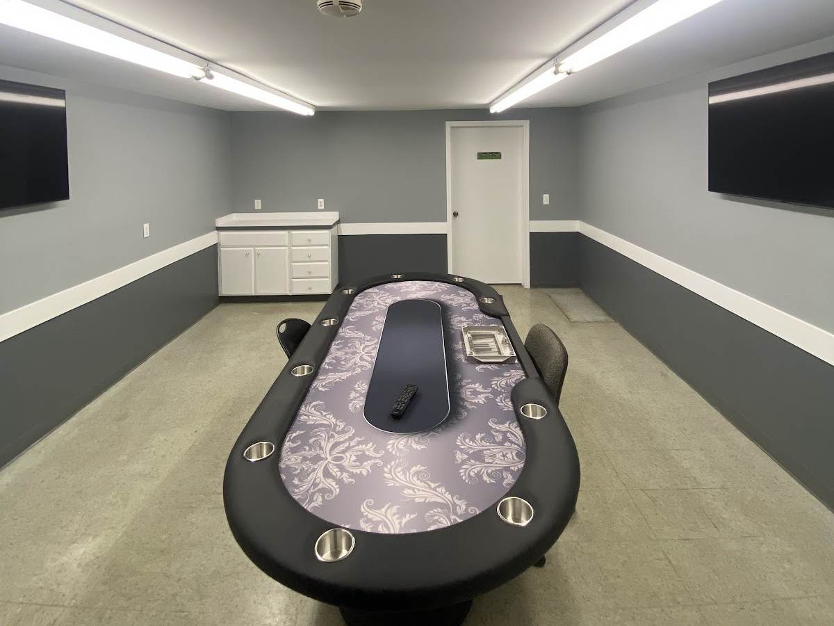 The same room painted light gray over a charcoal lower wall, with white trim, an uncovered poker table and two wall-mounted TVs.
