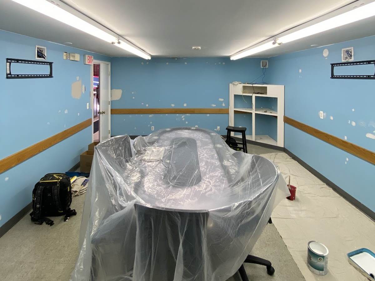 A long basement room with sky-blue walls dotted with patched spots, a brown chair rail and a poker table wrapped in plastic sheeting.