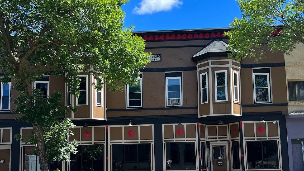 A two-story brick storefront painted in brown, black and cream trim with red diamond accents, a bay window above and leafy trees in front under a blue sky.