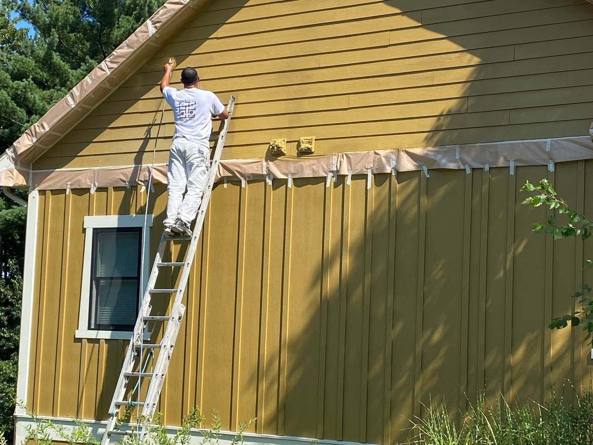 A painter on an aluminum ladder rolling paint onto a mustard-yellow board-and-batten wall with taped trim, a second crew member at the foot of the wall.