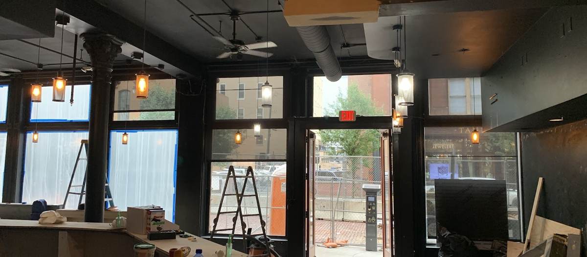 The inside of a storefront with black walls and ceiling, ladders, tools and drop cloths spread across the floor and a doorway to the street.